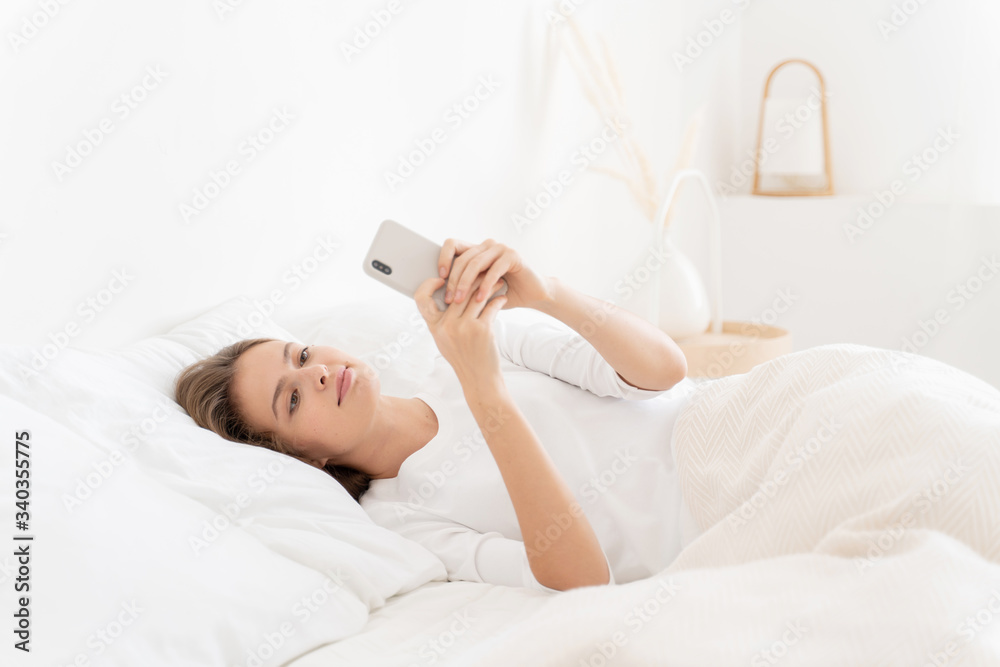 Young girl waking up in white bed late in morning, browsing Internet via her phone, answering text messages and enjoying free time Young girl waking up in white bed late in morning, browsing Internet via her phone, answering text messages and enjoying free time