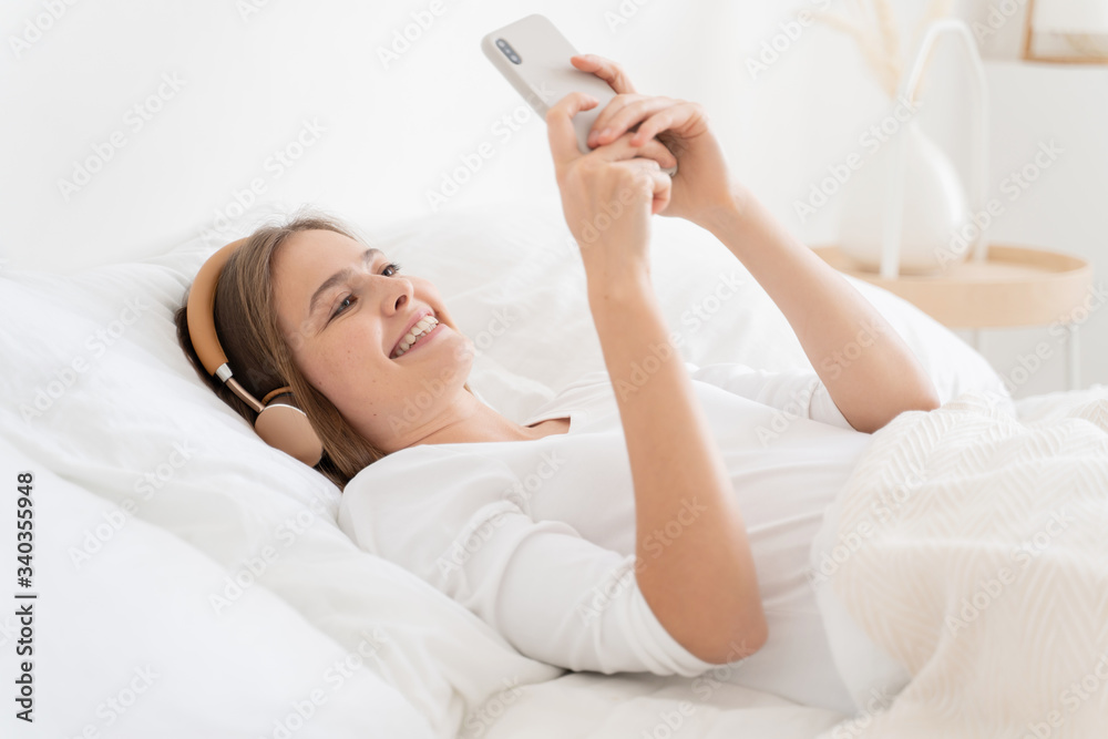 Young woman lying in white bed, dressed in top, having fun while listening to her friend through wireless headphones, enjoying video call on smartphone Young woman lying in white bed, dressed in top, having fun while listening to her friend through wireless headphones, enjoying video call on smartphone