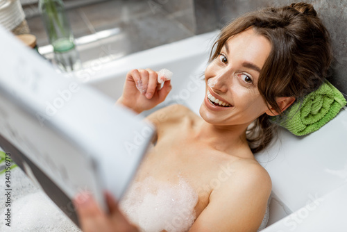 Portrait of a young relaxed woman taking a bath, lying in bathtube and reading some magazine at home
