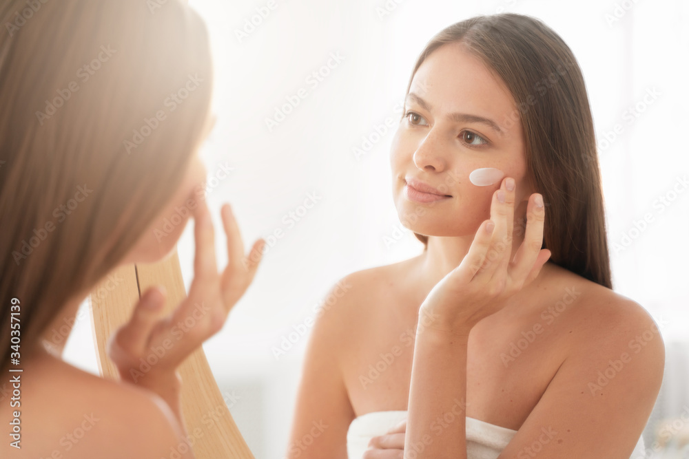 Young woman applying cream to her cheek in morning, looking in mirror in bathroom, standing wrapped in white bath towel, smiling Young woman applying cream to her cheek in morning, looking in mirror in bathroom, standing wrapped in white bath towel, smiling