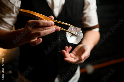 Close-up. Barman holding big cube of ice using tweezers over his other hand.