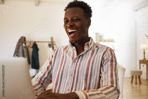 Happy cheerful young African male relaxing at home surfing internet on laptop, smiling broadly, looking for memes, playing vieo games or watching series online. Technology and leisure concept