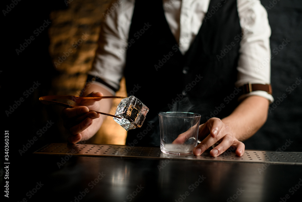 close-up. Bartender holds in hand tweezers with ice near steaming glass
