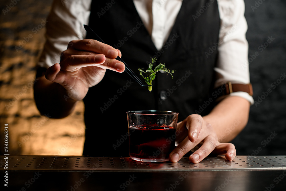 Close-up of glass with drink which bartender decorating with fresh green plant.