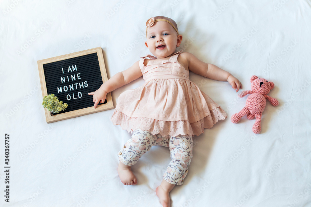 9 Nine months old baby girl laying down on white background with letter board and teddy bear. Flat lay composition. Stock Adobe Stock