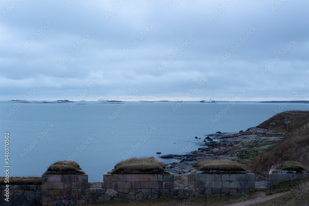 The rocky shore of Suomenlinna in the Gulf of Finland and the view of the lights of tourist ferries in autumn twilight in Finland. The harsh and laconic beauty of Finnish nature.