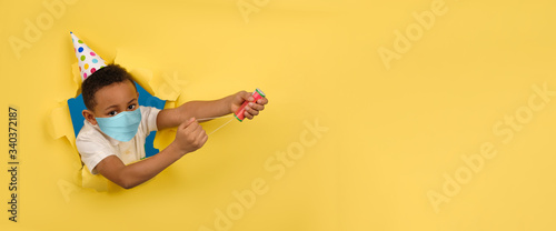 Sad African American boy celebrates his birthday by blowing up firecracker in medical mask protecting against coronavirus virus on yellow torn paper wall background. Concept of bad birthday.