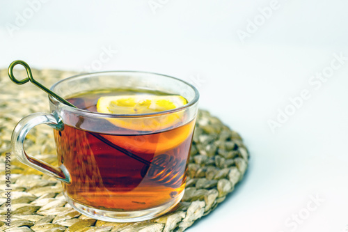 A transparent mug with black tea, a slice of lemon, a spoon for honey on a wicker gray napkin on a light table. The concept of a cozy tea party. Close-up