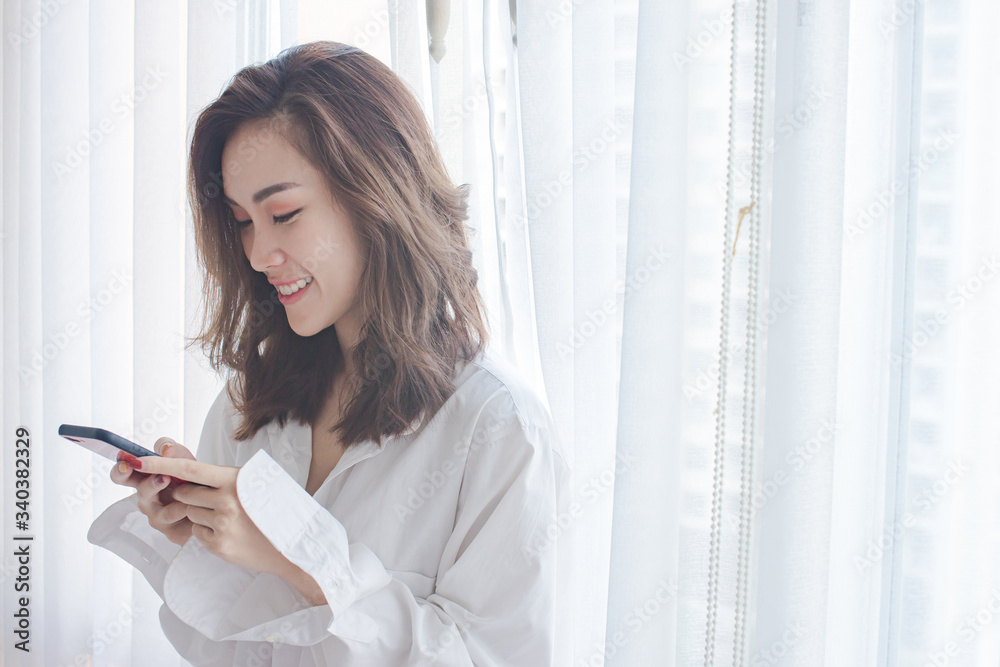 Attractive young woman chat on mobile phone and smiling while stand near window at home Attractive young woman chat on mobile phone and smiling while stand near window at home