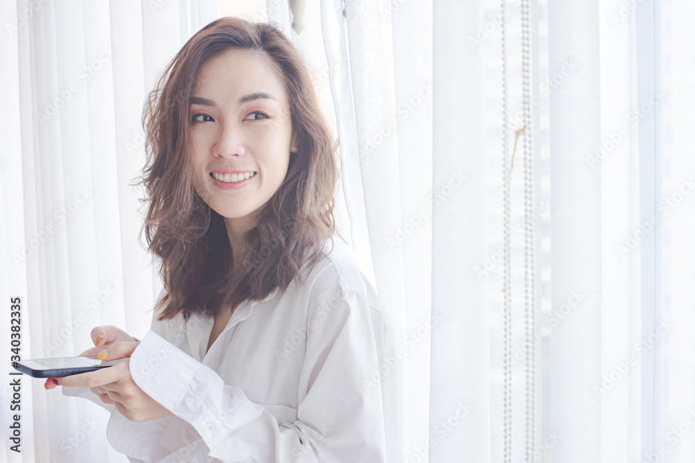 Attractive young woman chat on mobile phone and smiling while stand near window at home 