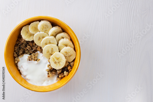 The muesli with banana and yogurt in a bowl on the table on a white wooden background.