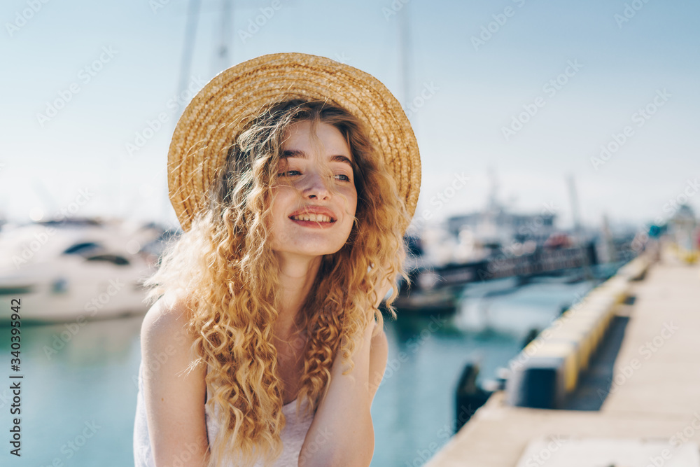 curly blonde in a straw hat sits on the background of the seaport and gently smiles