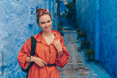 the blue city of morocco behind the back of a European girl in a bright orange dress with a backpack behind her shoulders and sunglasses on her head © nelen.ru