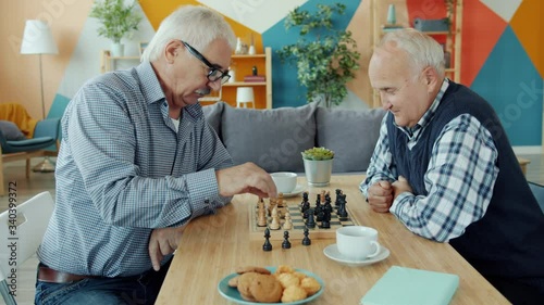 Slow motion of male friends playing chess and talking sitting at table indoors in apartment. Retirement, friendhsip and leisure activities concept.