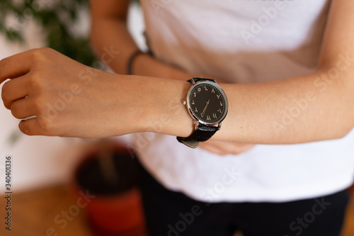 close up photo of female hand wearing black wrist watch with arabic numerals with green flower in background.