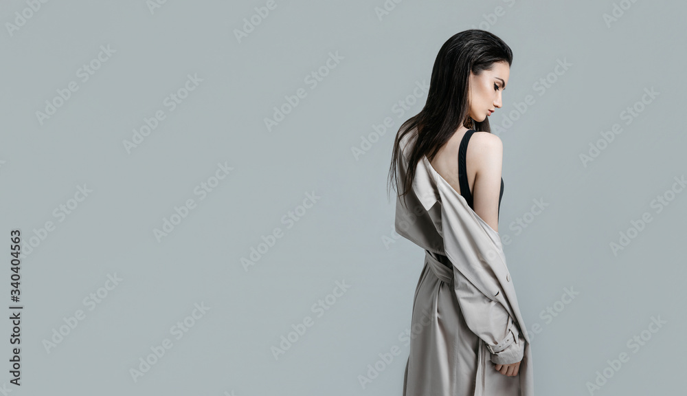 Fashion portrait of a young sexy stylish brunette woman in black bodysuit, beige trench coat on a gray background in the studio. Model with perfect skin and professional liner make-up Fashion portrait of a young sexy stylish brunette woman in black bodysuit, beige trench coat on a gray background in the studio. Model with perfect skin and professional liner make-up