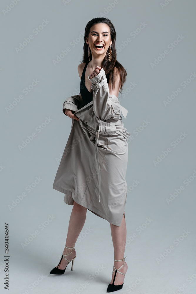 Smiling cheerful joyful girl on heels in a black bodysuit, beige trench coat with large earrings posing on gray background in the studio. Full-length portrait of a girl with straight dark hair Smiling cheerful joyful girl on heels in a black bodysuit, beige trench coat with large earrings posing on gray background in the studio. Full-length portrait of a girl with straight dark hair