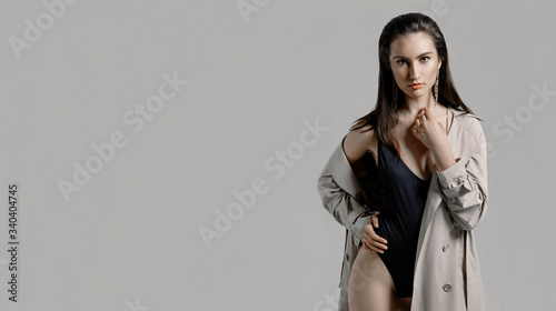 Fashion portrait of a young sexy stylish brunette woman in black bodysuit, beige trench coat on a gray background in the studio. Model with perfect skin and professional liner make-up