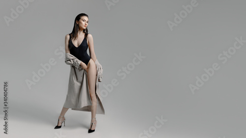 Beautiful young sexy brunette woman on heels in a black bodysuit, beige trench coat with large earrings posing on gray background in the studio. Full-length portrait of a girl with straight dark hair