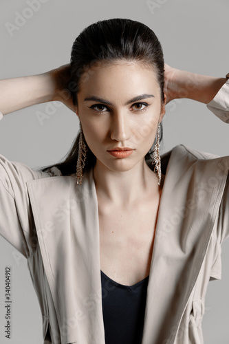 Beautiful sexy brunette woman in a black bodysuit, a beige trench coat with large gold earrings posing on a gray background in the studio. Close-up portrait of a model with professional liner make-up 