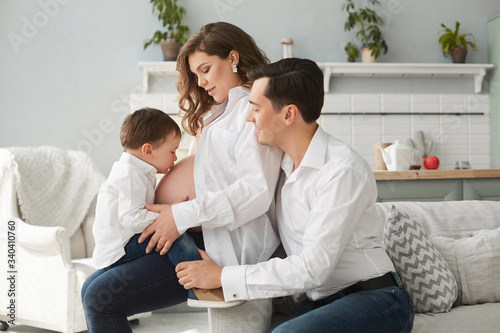 Side view of cute pregnant woman resting with family at home. Little boy kissing belly of mother while happy father sitting near and hugging wife. Cheerful family in white shirts. Expecting baby