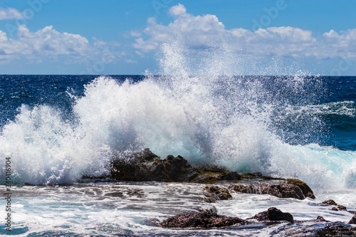 Wave breaking on rock offshore Hawaii's Big Island. Deep blue pacific ocean, sky and clouds in the distance. 