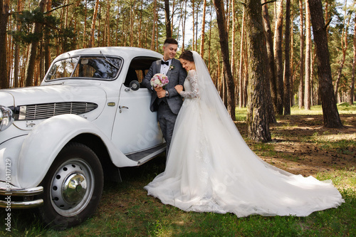 Beautiful bride in luxury wedding dress with long train and veil and elegant groom in grey suit posing near white Just Married car. Stylish wedding couple, elegant newlyweds