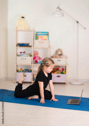Little girl in sportswear watching online video on laptop and doing workout fitness exercises in her room at home. Distant online training, social distance or self-isolation, online education concept.