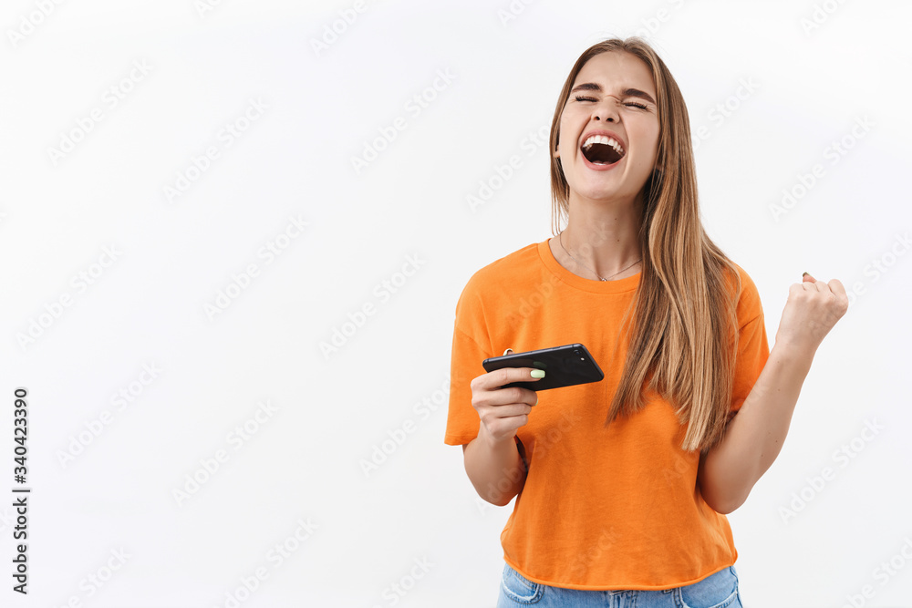 Technology, youth and communication concept. Triumphing, happy lucky blonde girl winning in game, upload cool racing or shooter on mobile phone, fist pump shouting yes with closed eyes and smile Technology, youth and communication concept. Triumphing, happy lucky blonde girl winning in game, upload cool racing or shooter on mobile phone, fist pump shouting yes with closed eyes and smile