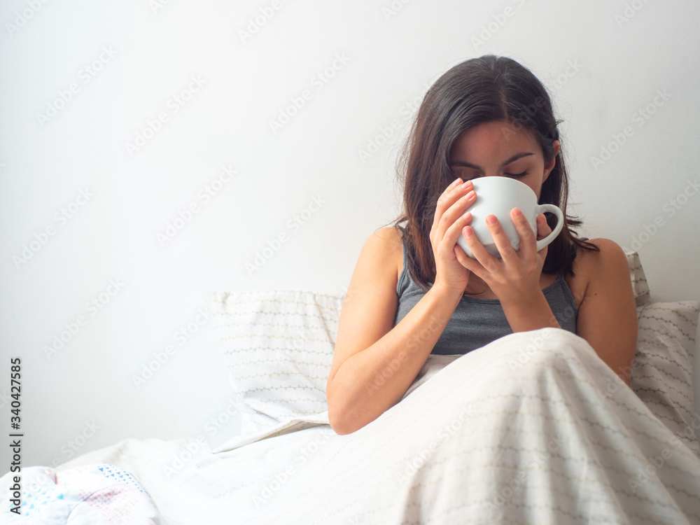 Young Brown Hair Hispanic Female Drinks Tea or Coffee in a White Cup in her Bed in Quarantine Young Brown Hair Hispanic Female Drinks Tea or Coffee in a White Cup in her Bed in Quarantine