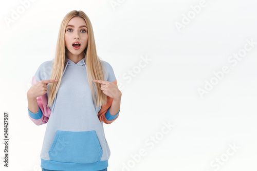 Curious excited blonde girl, teenage college student asking permition to participate, pointing fingers at herself, open mouth look camera curious, show-off talk personal achievement, white background photo