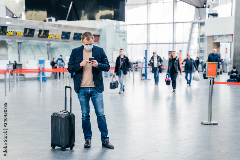 Horizontal shot of man poses in crowded airport, uses cell phone, checks time of flight online, stands near suitcase, wears medical mask during coronavirus crisis. Health care, traveling concept