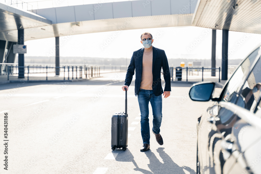 Business traveler pulls suitcase from modern airport terminal, walks to car, wears medical disposable mask on face, prevents from coronavirus during dangerous pandemic situation. Full length shot Business traveler pulls suitcase from modern airport terminal, walks to car, wears medical disposable mask on face, prevents from coronavirus during dangerous pandemic situation. Full length shot