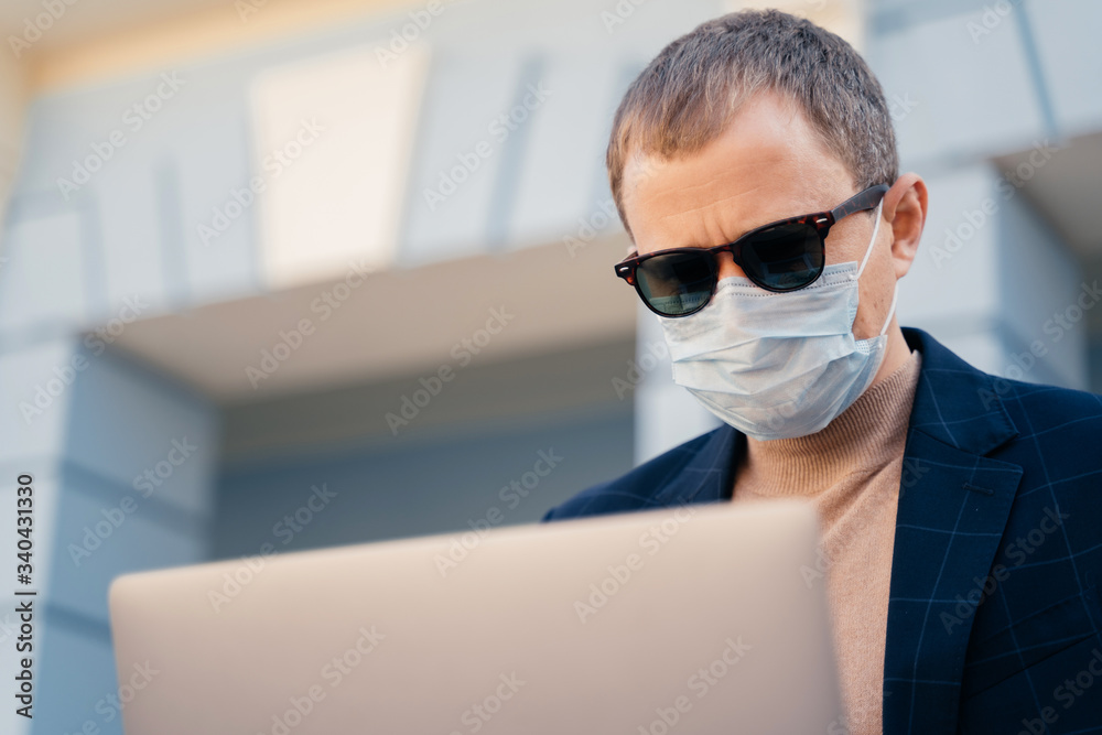 Close up shot of adult man wears sunglasses and medical mask, reads news  online on laptop computer, finds out symptoms of coronavirus, has online communication  during quarantine and self isolation Stock Photo