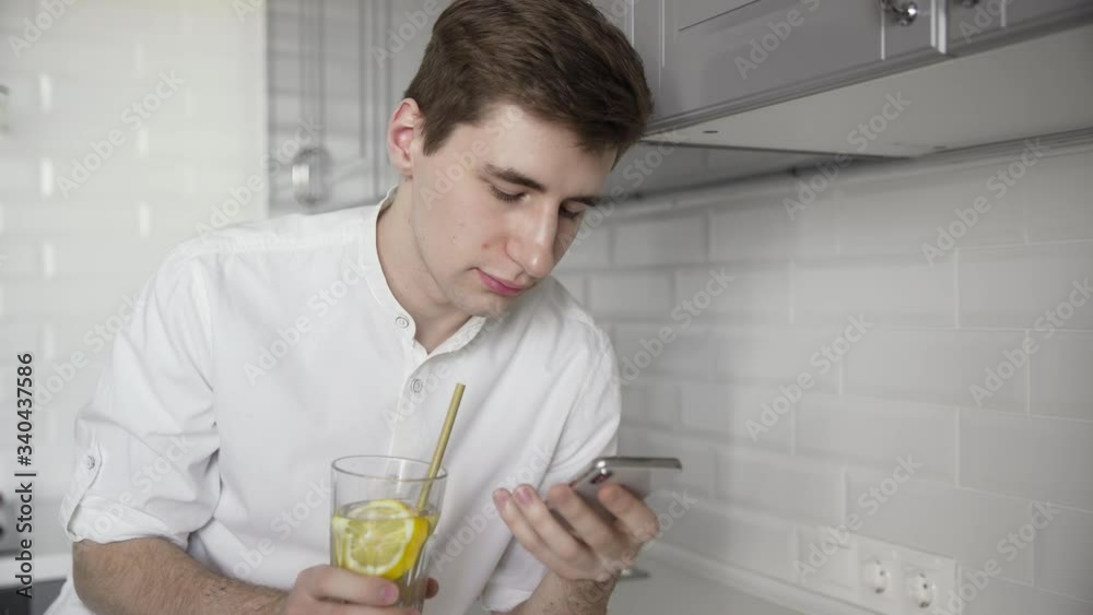 Young man in a white short looks at the smartphone and uses reusable eco-friendly ecological bamboo straw to drink water with lemon at the kitchen