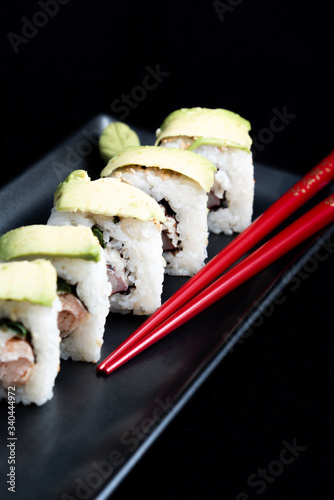 Sushi roll with avocado wrap, grilled salmon, Philadelphia cheese and green onions on black plate and red chopsticks. Small pot with soy sauce. Black background.