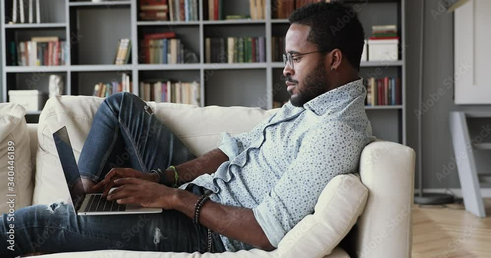Side view young smart multiracial guy in eyeglasses leaning on comfortable sofa, working remotely on computer from home. Focused millennial african ethnic student studying distantly on online courses.