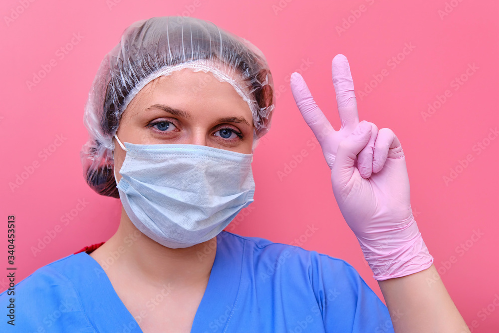 A female doctor in a medical mask on a red background shows a victory gesture. A medic with an approving hand sign, the concept of defeating the coronavirus epidemic. A female doctor in a medical mask on a red background shows a victory gesture. A medic with an approving hand sign, the concept of defeating the coronavirus epidemic.
