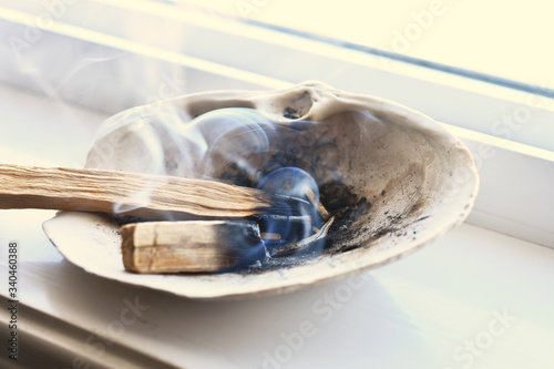 An image of a burning palo santo smudge stick in a seashell on a white window ledge. 