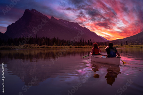 Couple adventurous friends are canoeing in a lake surrounded by the Canadian Mountains. Dramatic Sunset Composite. Taken in Vermilion Lakes, Banff, Alberta, Canada.