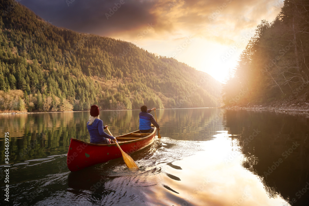 Couple friends canoeing on a wooden canoe during a colorful sunny sunset. Cloudy Sky Composite. Taken in Harrison River, East of Vancouver, British Columbia, Canada.