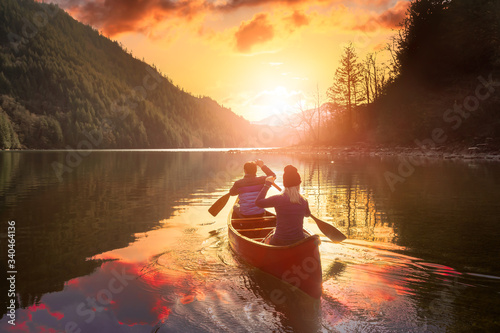 Couple friends canoeing on a wooden canoe during a colorful sunny sunset. Cloudy Sky Composite. Taken in Harrison River, East of Vancouver, British Columbia, Canada.