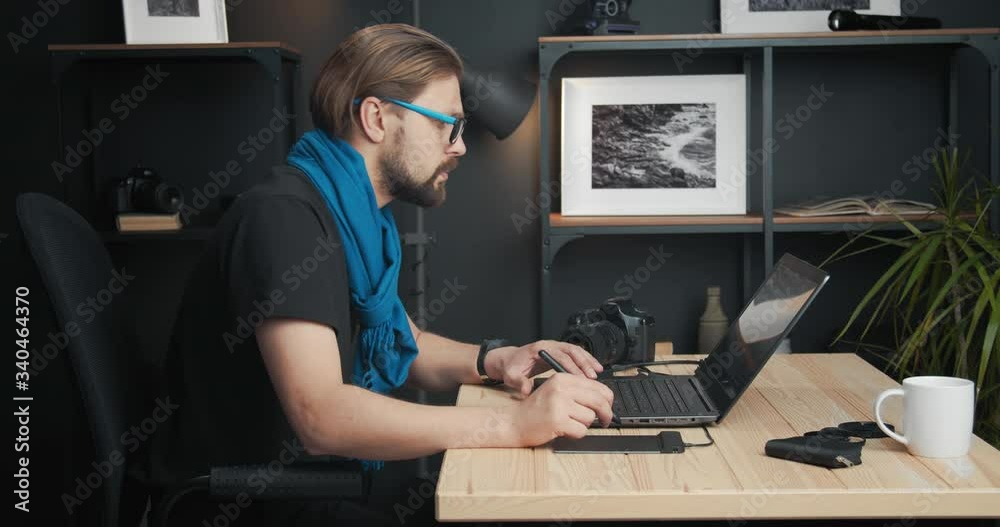 Side view of bearded photographer in eyeglasses drawing and retouching images on laptop using stylus pen. Mature man in black t-shirt and blue scarf working at modern studio.