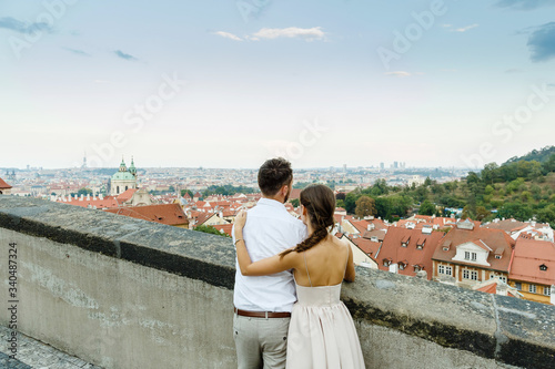 Rear view of young romantic couple of groom and bride standing on the rooftop and enjoying beautiful view of the city Prague, Czech Republic