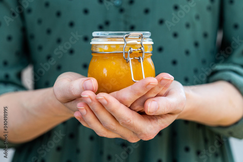 Female hands holding a home made vegan raw apricot jam in a glass jar with a olive green dress. Healthy raw yellow jam in a cup.
