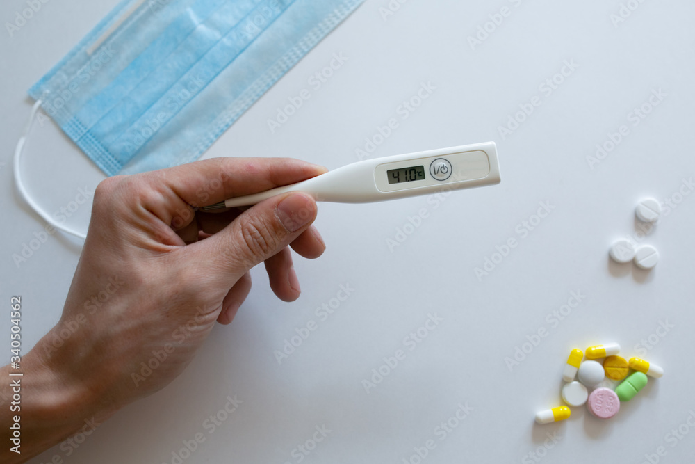 In the hands of the doctor is an electronic thermometer for measuring temperature, a protective mask from coronavirus and tablets, medications on a blue background .COVID-2019. In the hands of the doctor is an electronic thermometer for measuring temperature, a protective mask from coronavirus and tablets, medications on a blue background .COVID-2019.