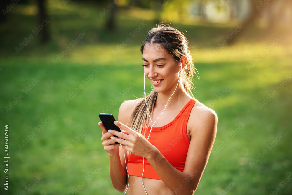 Portrait of young slim charming sportswoman standing in nature, having earphones in ears and using smart phone for choosing music.