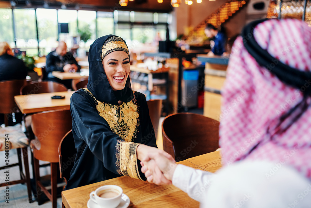 Cute smiling positive arab woman dressed in traditional wear shaking hands with her male business partner. Coffee shop interior. Diversity concept.