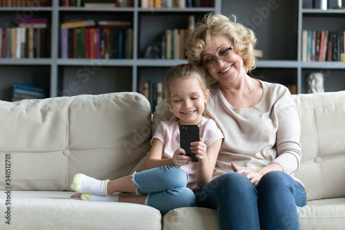 Multi generational family and modern tech, parental control, children protection in internet concept. Grandma sit on sofa with granddaughter holds cellphone use new app for kids, read e-books on-line