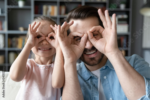 Small daughter and father have fun at home, do funny faces making with fingers eyewear shape like glasses looking at camera through binoculars, eyeglasses sunglasses lenses store advertisement concept